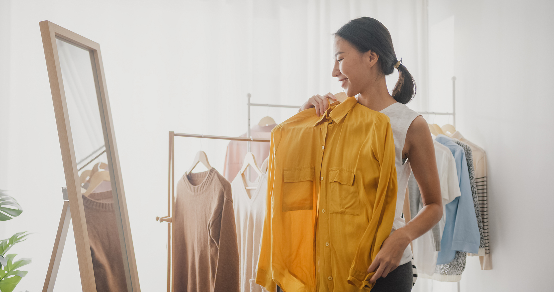 Young Asian woman choosing clothes on clothes rack dressing looking herself in mirror in living room at home.